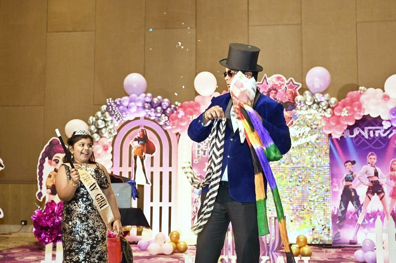A magician pulling rainbow silks from a newspaper as the birthday girl watches